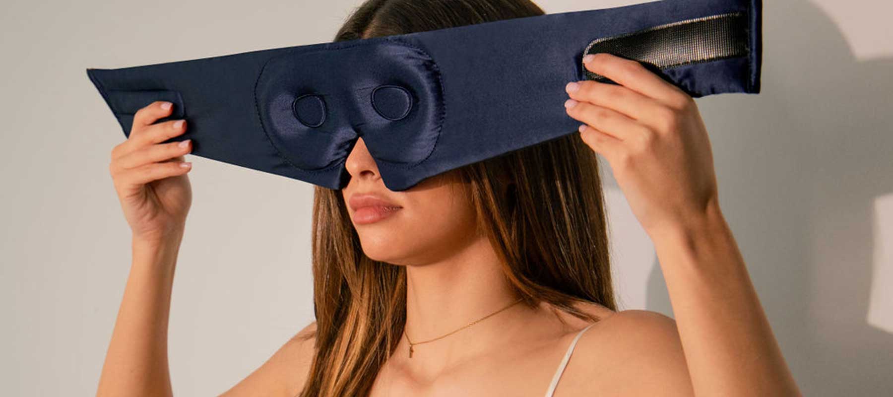 A woman with long brown hair holds a large, padded navy blue eye mask with Velcro straps up to her face, preparing to put it on. She wears a thin-strapped top and a delicate necklace.