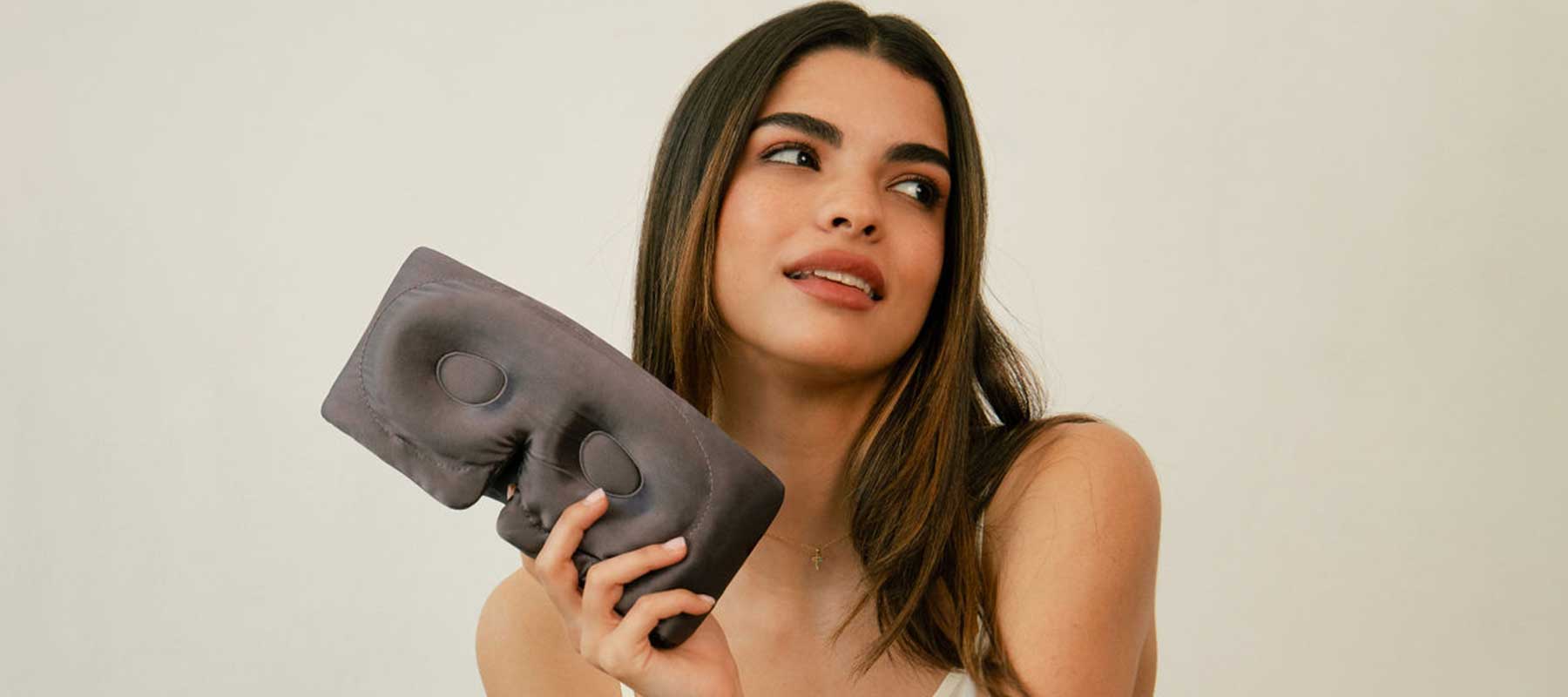 A young woman with long dark hair holds a gray sleep mask and looks off to the side, smiling slightly, against a plain light background.