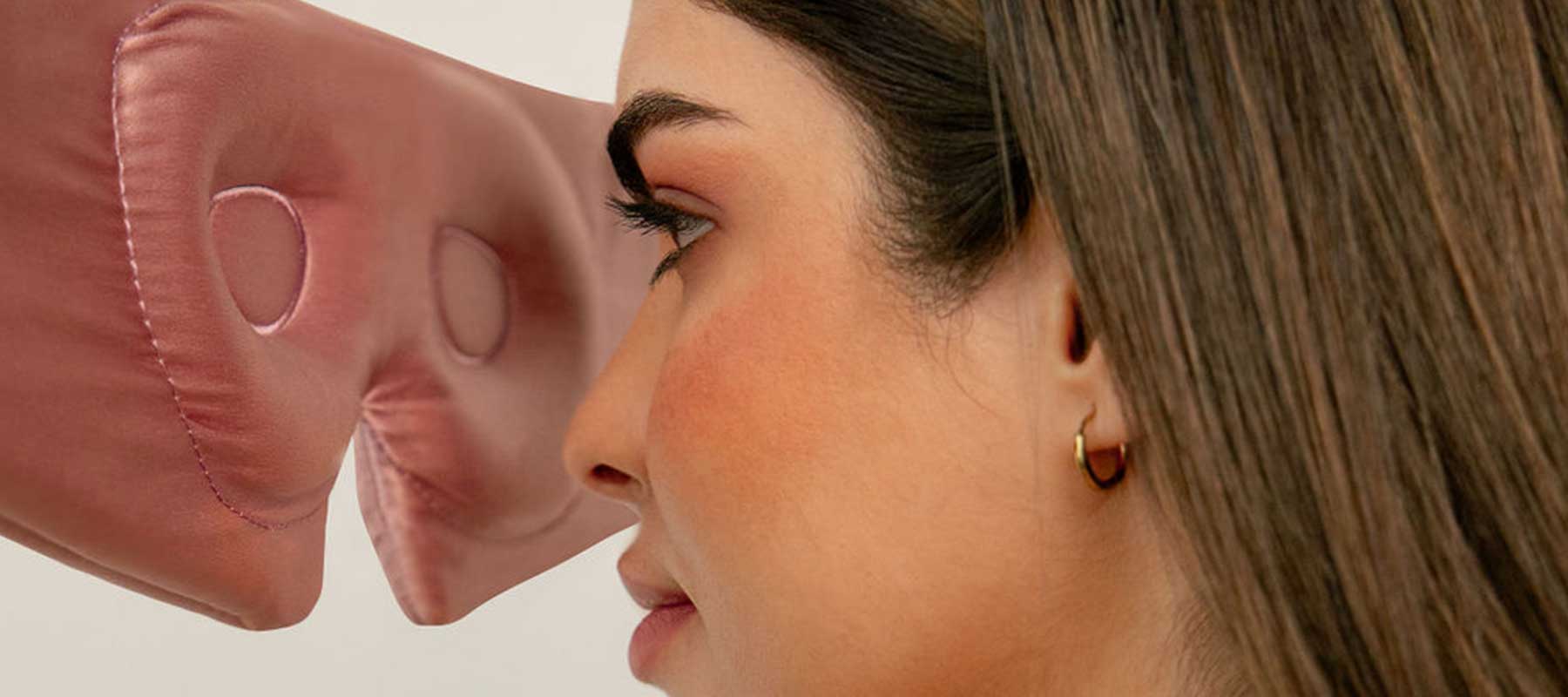 A woman with brown hair and gold hoop earrings faces a large, pink, inflatable nose in front of her against a neutral background.