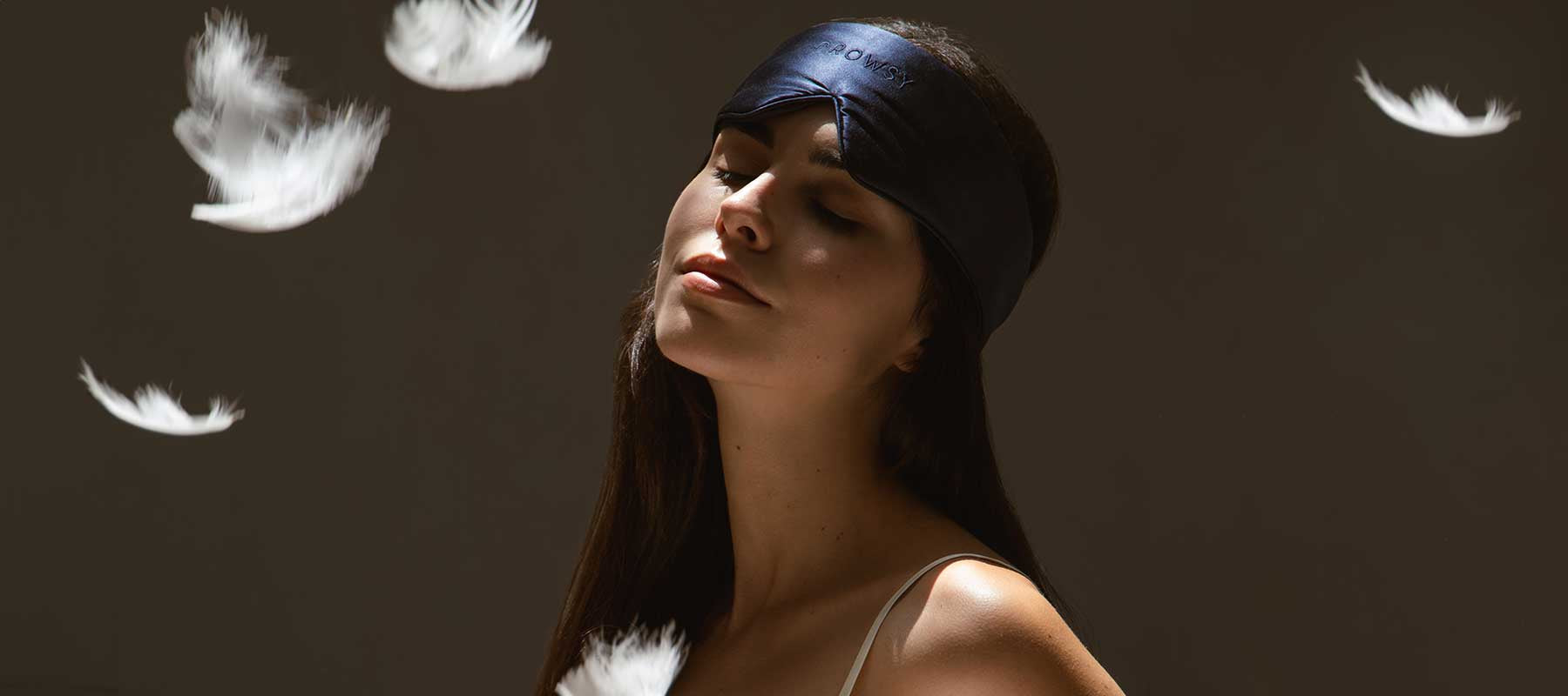 A woman wearing a black sleep mask tilts her head slightly back with a peaceful expression, surrounded by floating white feathers against a dark background.