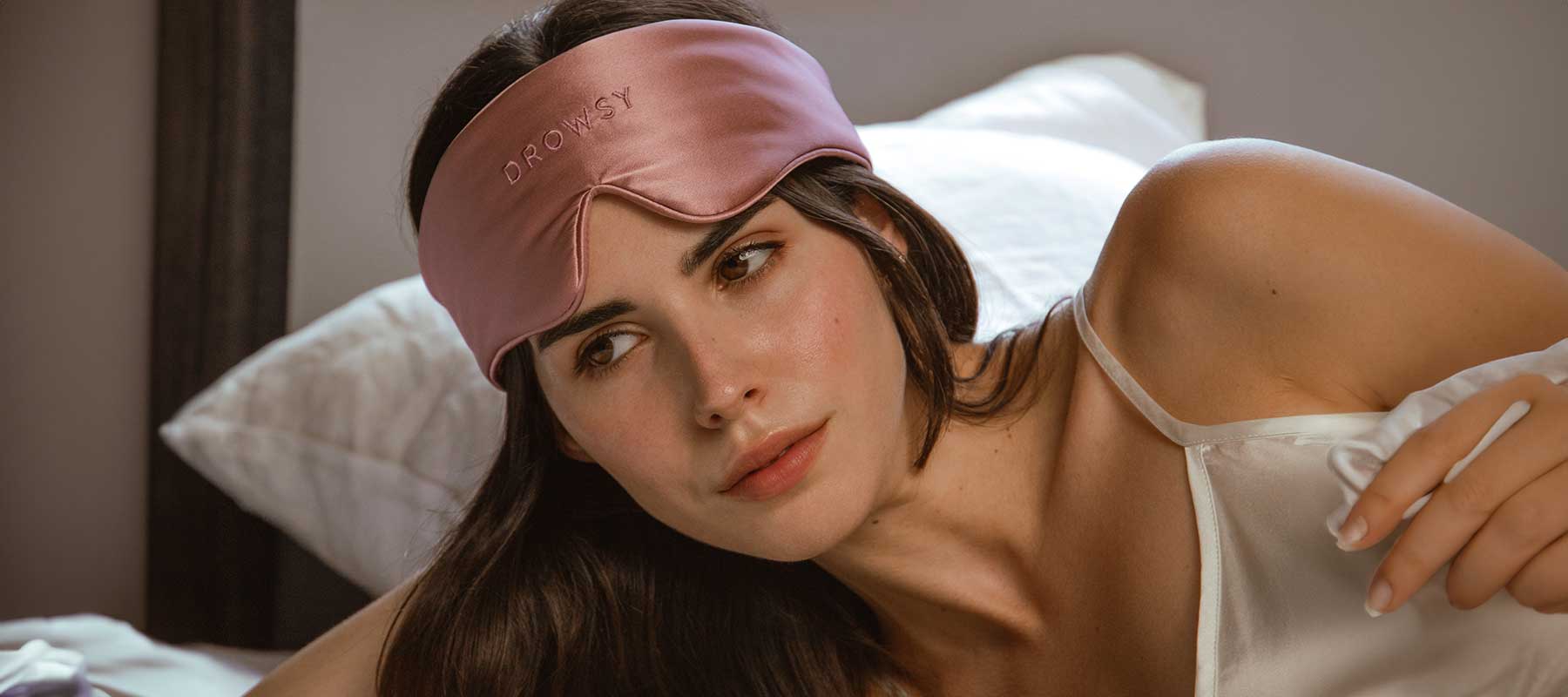 A woman with dark hair lies on a bed wearing a pink sleep mask pushed up on her forehead. She is dressed in a white satin top and rests on white pillows, looking thoughtfully to the side.