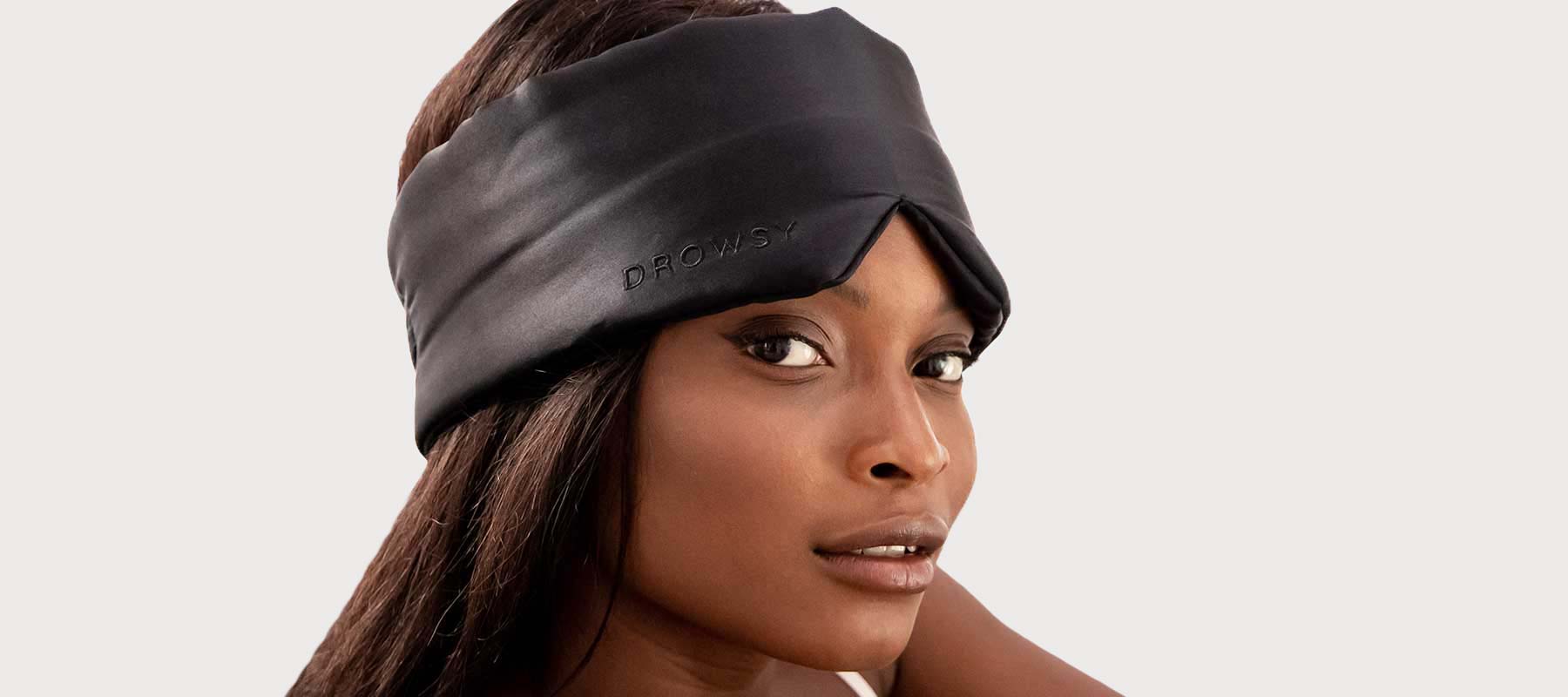 A woman with long dark hair wears a wide, black silk sleep mask on her forehead, looking confidently at the camera against a plain, light background.
