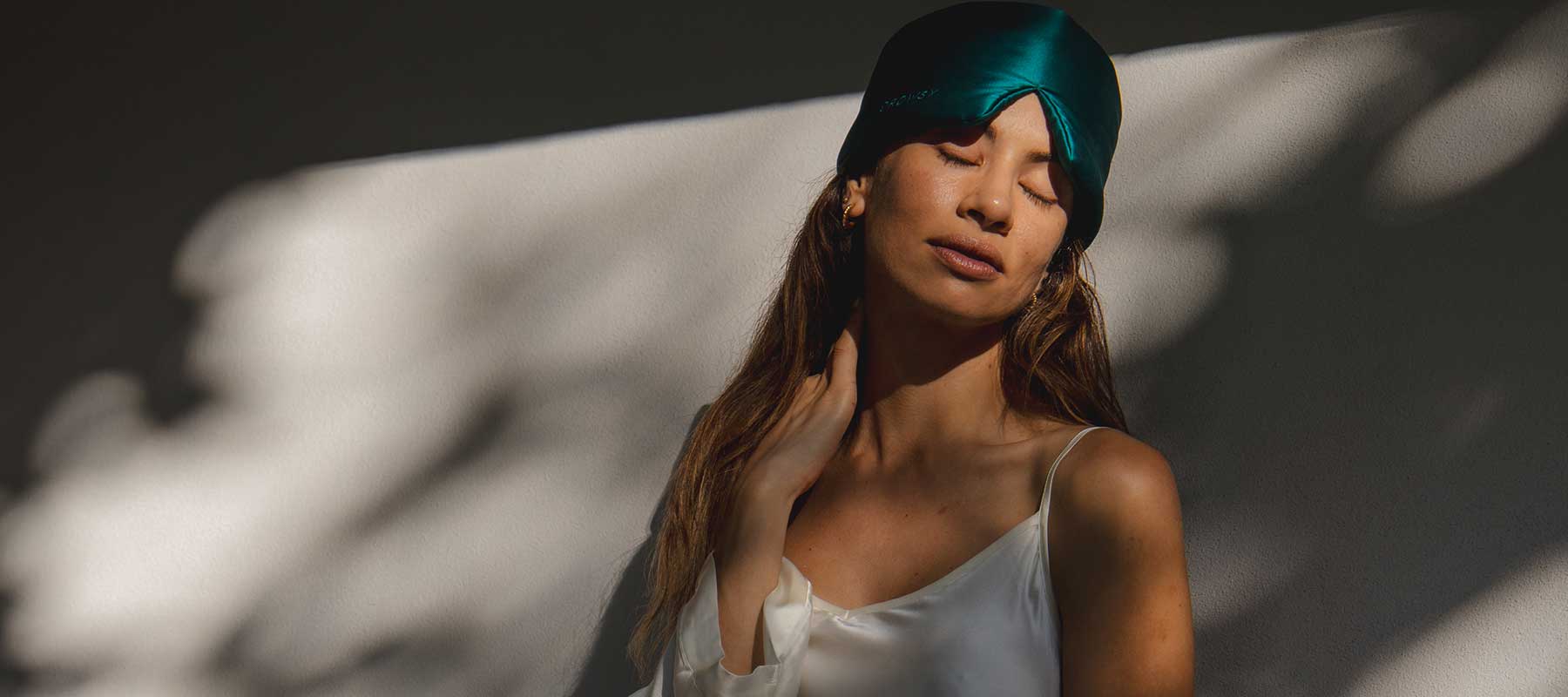 A woman wearing a teal satin sleep mask on her head and a white camisole stands against a sunlit wall with shadows, eyes closed and hand gently touching her neck.