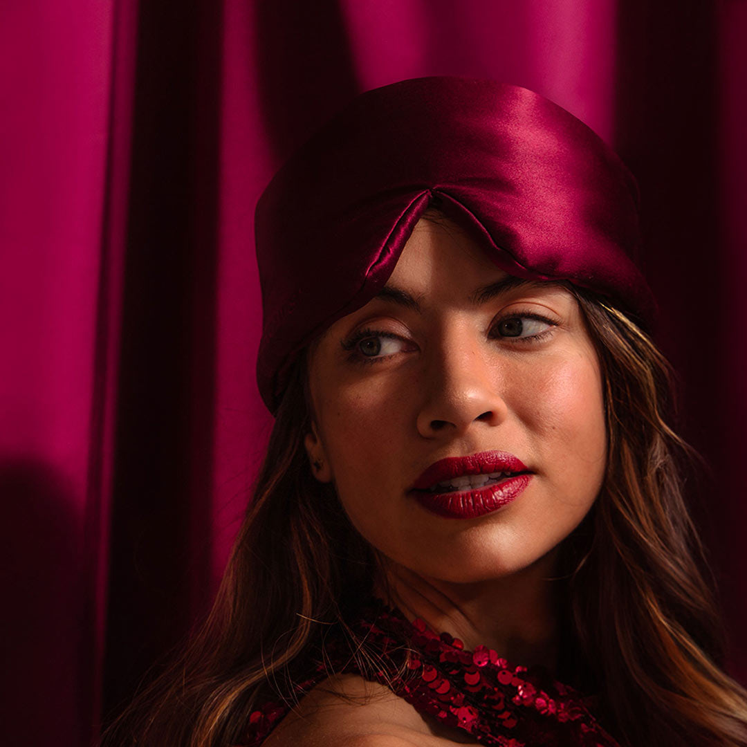 A woman wearing a shiny, deep red headscarf and matching lipstick looks to the side against a background of rich red curtains. She is dressed in a red sequin top.