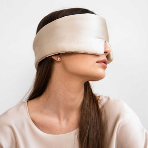 A woman with long brown hair models the drowsysleepco Sensual Sleep Bundle in Dusty Gold, featuring a beige silk sleep mask and matching satin top, posed against a plain white background.