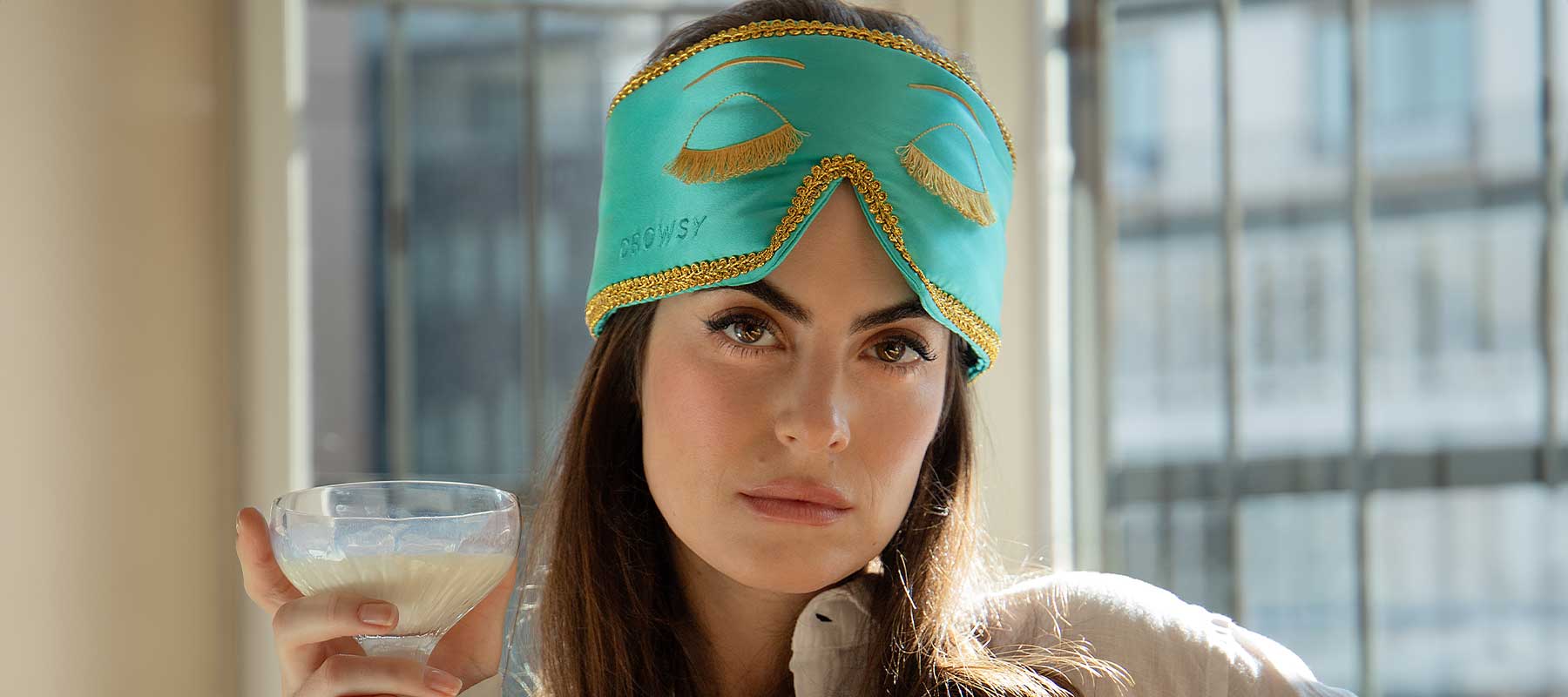 A woman with long brown hair wears a turquoise sleep mask with gold trim and eyelash designs pushed up on her forehead, holding a glass of a creamy beverage, standing indoors in front of large windows with bars.