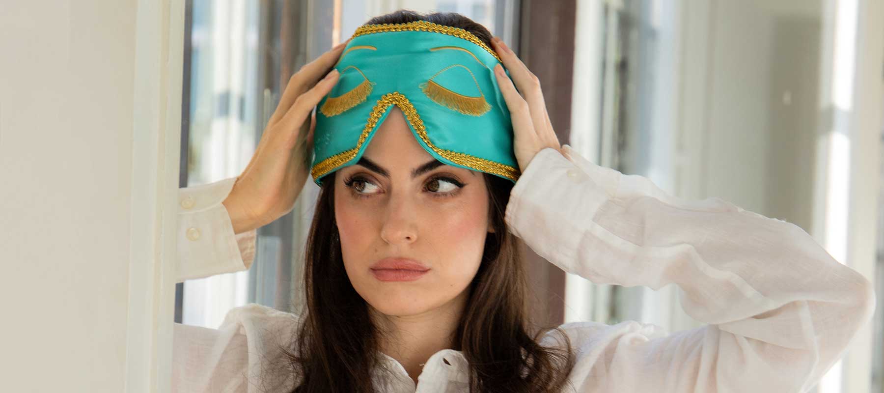 A woman wearing a white blouse adjusts a turquoise sleep mask with gold trim on her head, looking thoughtfully to the side in a bright room.