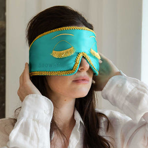 A woman in a white shirt adjusts the drowsysleepco Perfect Skin Bundle "Breakfast at Drowsy" turquoise silk sleep mask with gold trim and lash embroidery over her eyes, standing indoors against a light background.