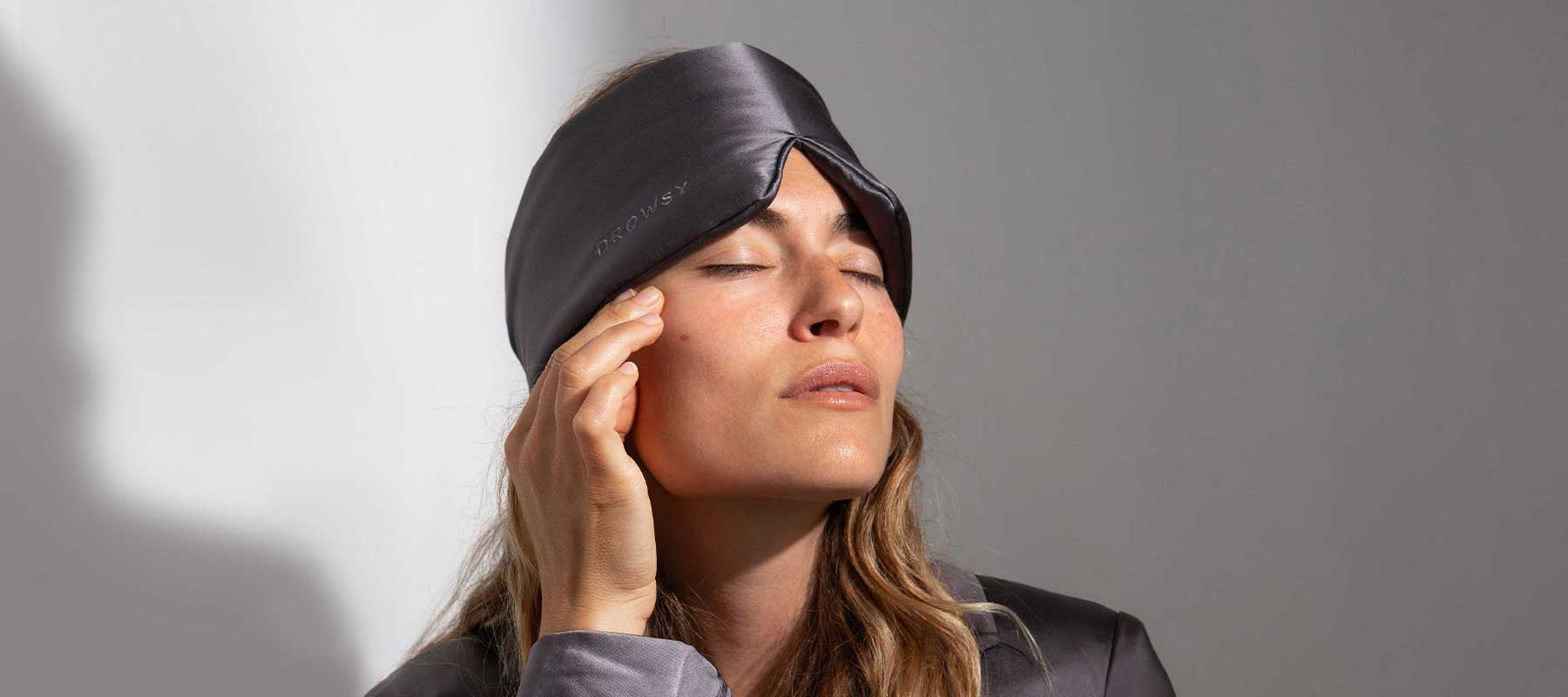 A woman with long hair wears a gray silk sleep mask on her forehead and lightly touches her temple with her fingers, eyes closed, appearing relaxed. She is dressed in matching gray sleepwear.
