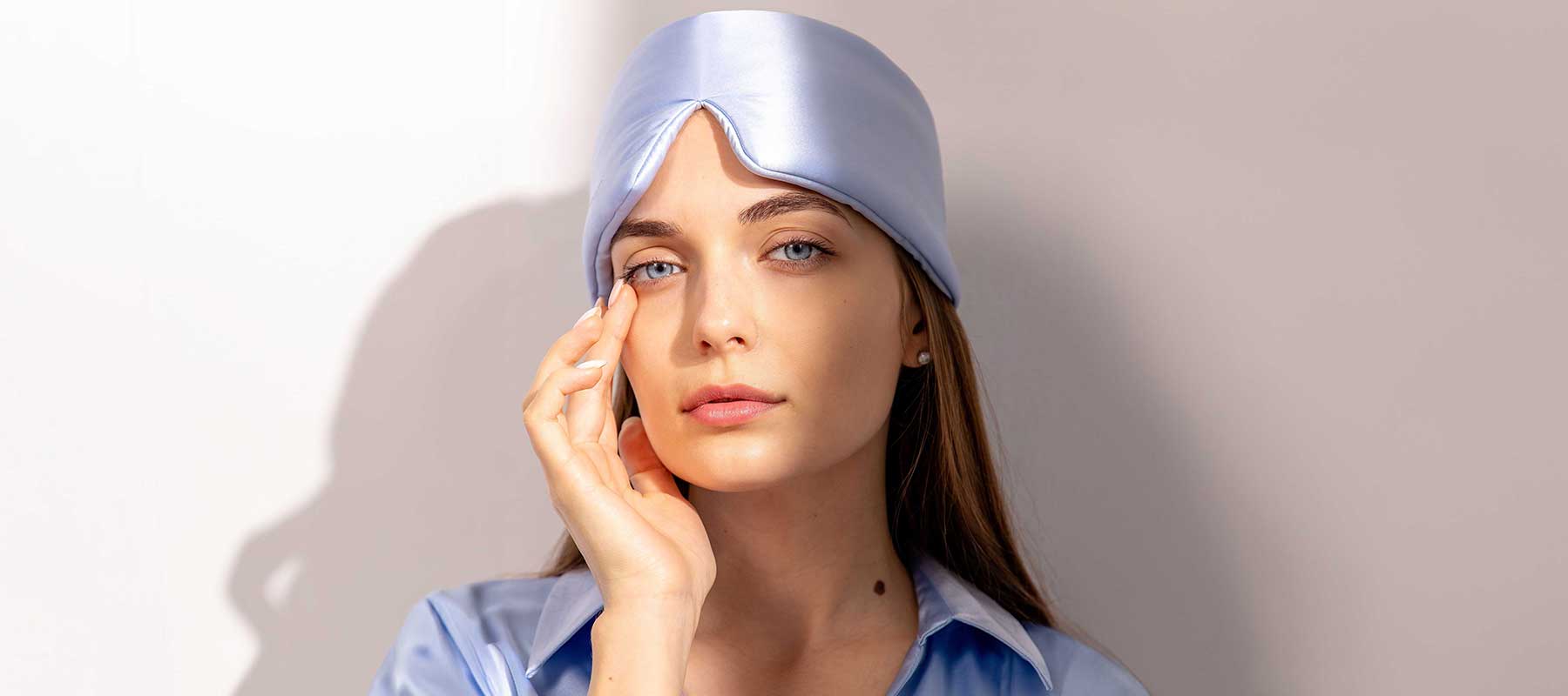 A woman with long hair wears a shiny, light blue sleep mask lifted onto her forehead and a matching satin shirt, gently touching her face with one hand while looking at the camera against a plain background.