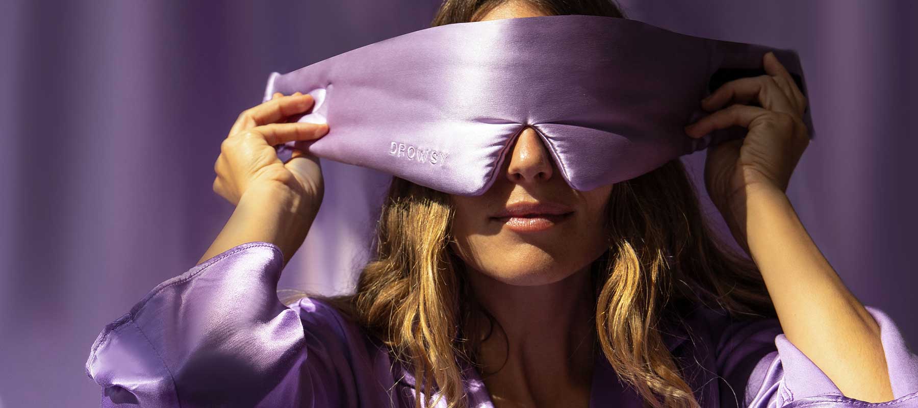 A person in a shiny lavender robe adjusts a large, silky purple sleep mask over their eyes against a matching purple background.