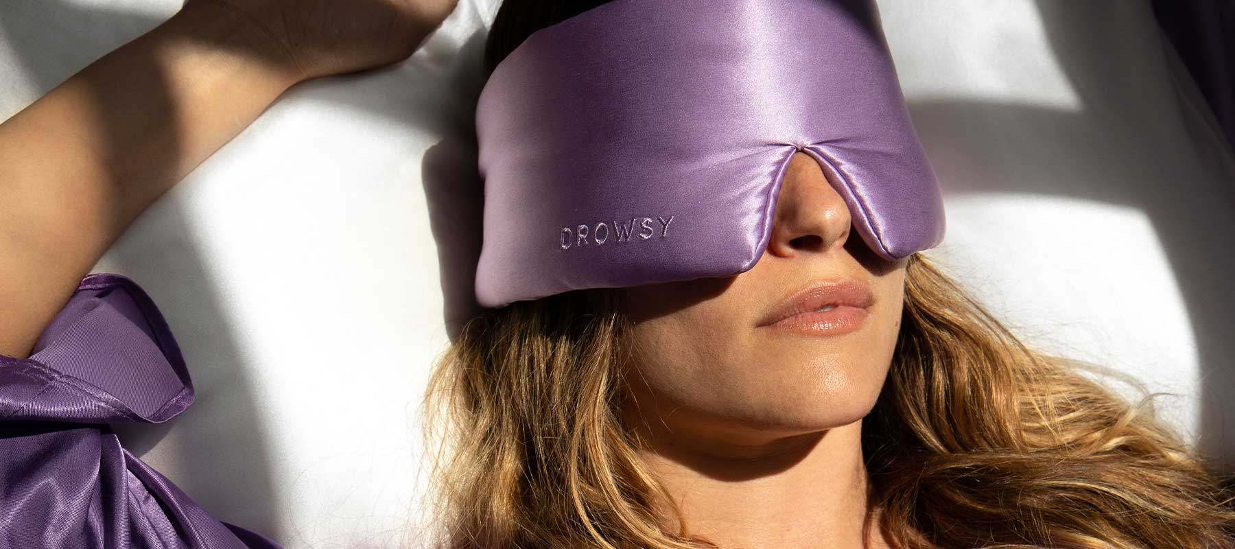 A person with wavy hair is lying down, wearing a shiny lavender eye mask labeled DROWSY and a matching robe, resting in bright sunlight.