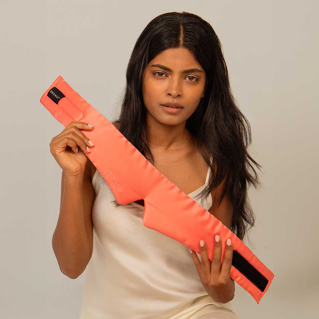 A woman with long dark hair wearing a white sleeveless top holds a bright orange, rectangular wrap with a black strap and the word thermotex printed on it. She looks at the camera against a plain background.