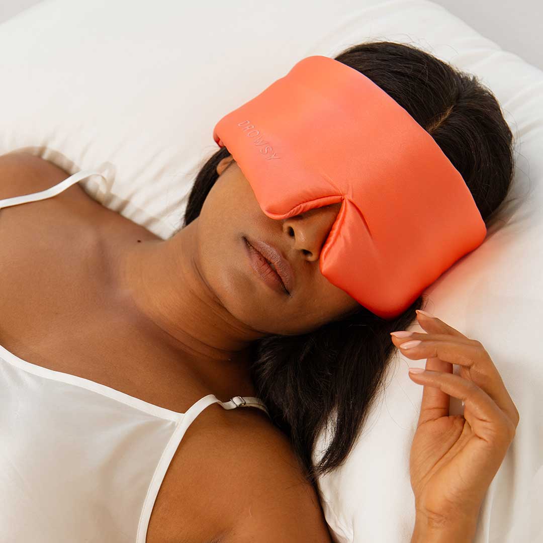 A woman wearing a white camisole lies on a bed with a white pillow, using a large orange sleep mask that covers her eyes and part of her face.