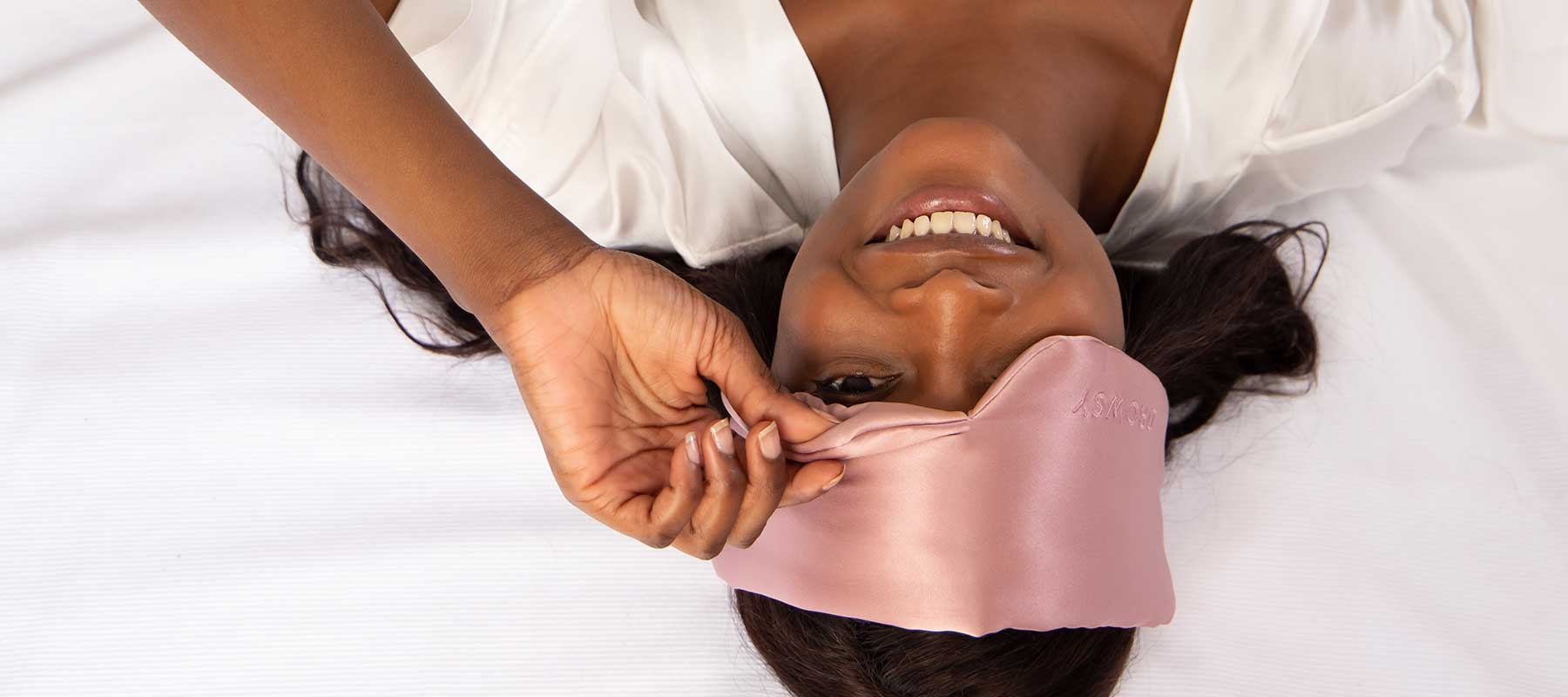 A woman lying on a bed in a white robe, smiling as she adjusts a pink satin sleep mask on her forehead.