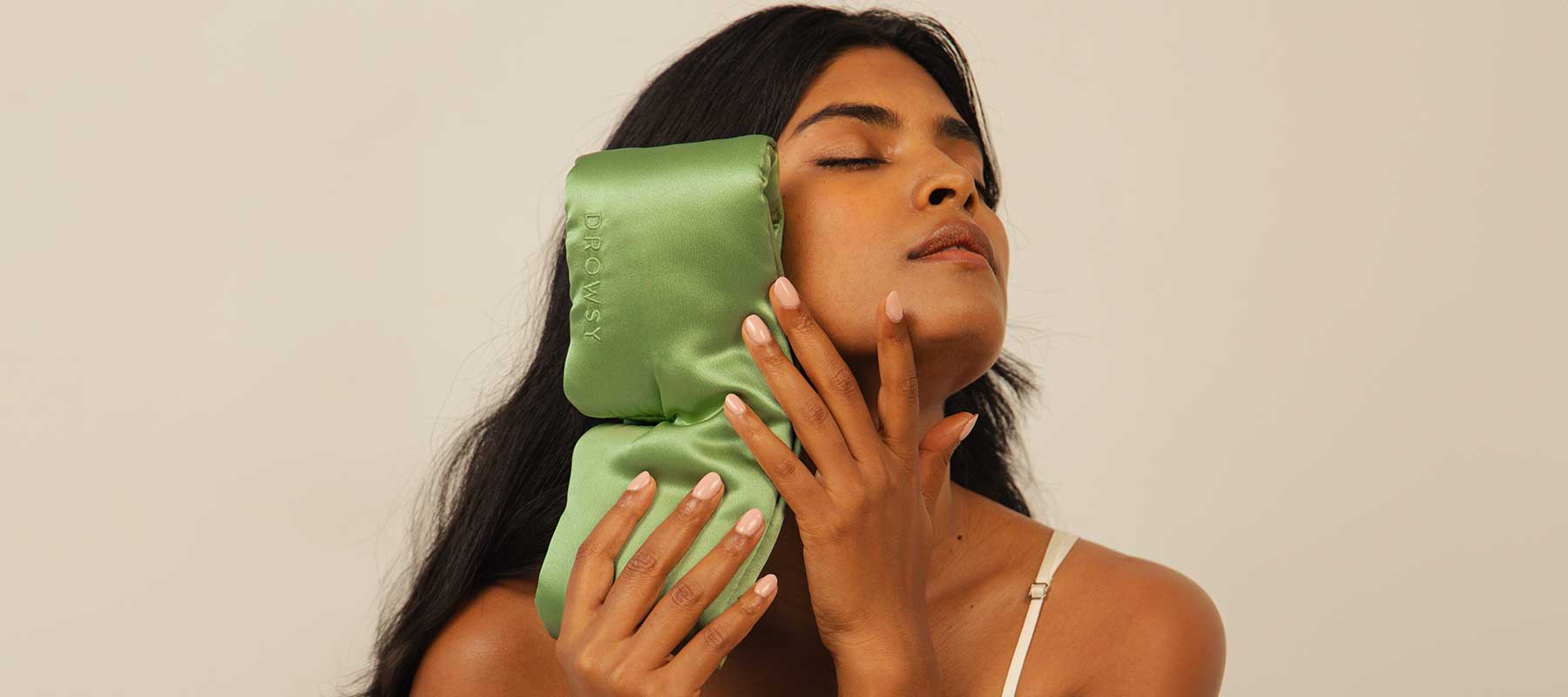 A woman with long dark hair holds a green satin pillow or ice pack to the side of her face, eyes closed, wearing a cream-colored camisole against a neutral background.