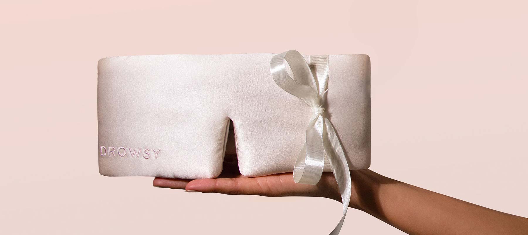 A hand holds a pale pink silk sleep mask with a ribbon tie and the word Drowsy embroidered in the corner, against a soft, light pink background.