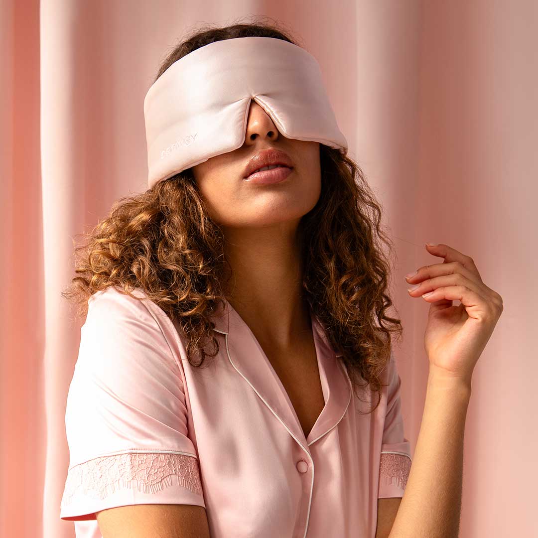A woman with curly hair wearing light pink pajamas sits against a pink background, with a satin eye mask covering her eyes. She has a relaxed expression and her hand is gently raised beside her face.