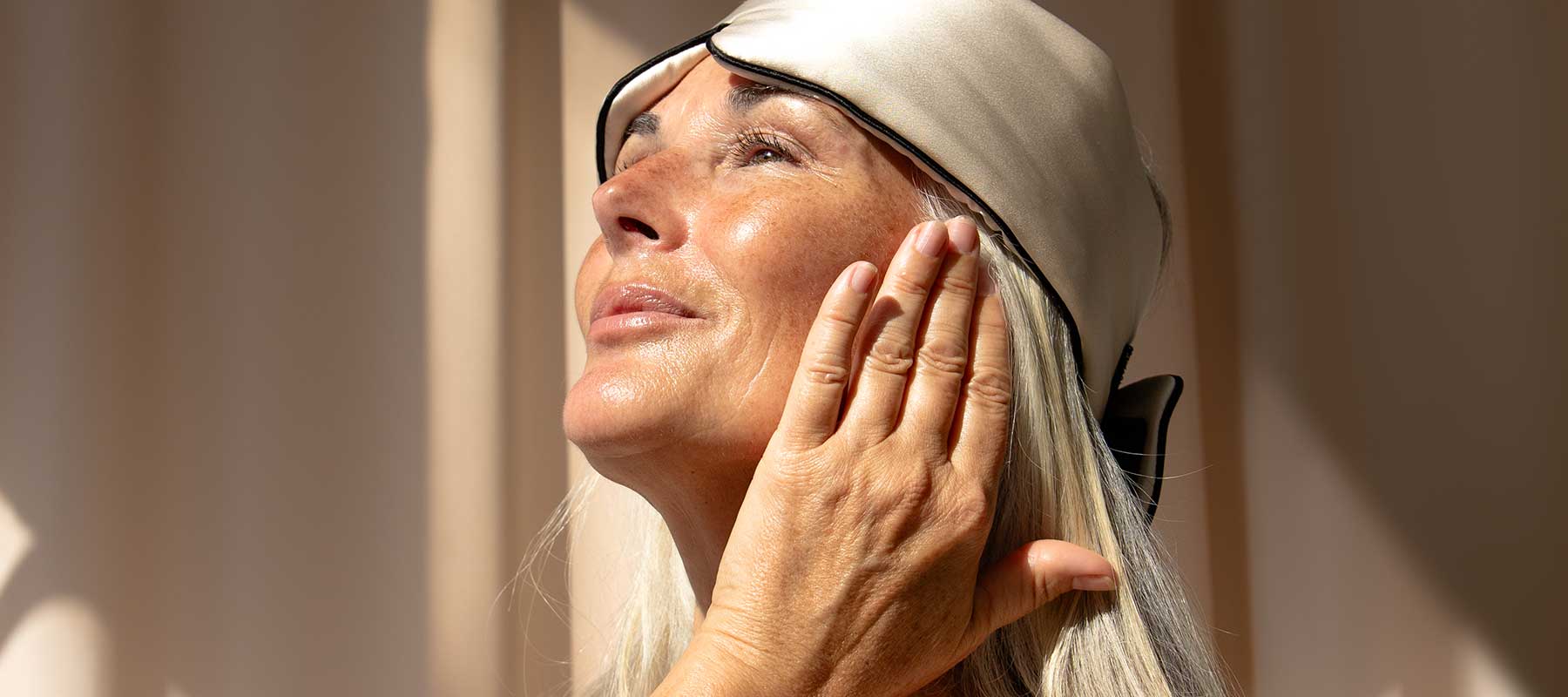 A mature woman with long gray hair and a satin sleep mask on her forehead gently touches her face, looking up toward the sunlight with a peaceful expression. Soft light and shadows fall across her face.