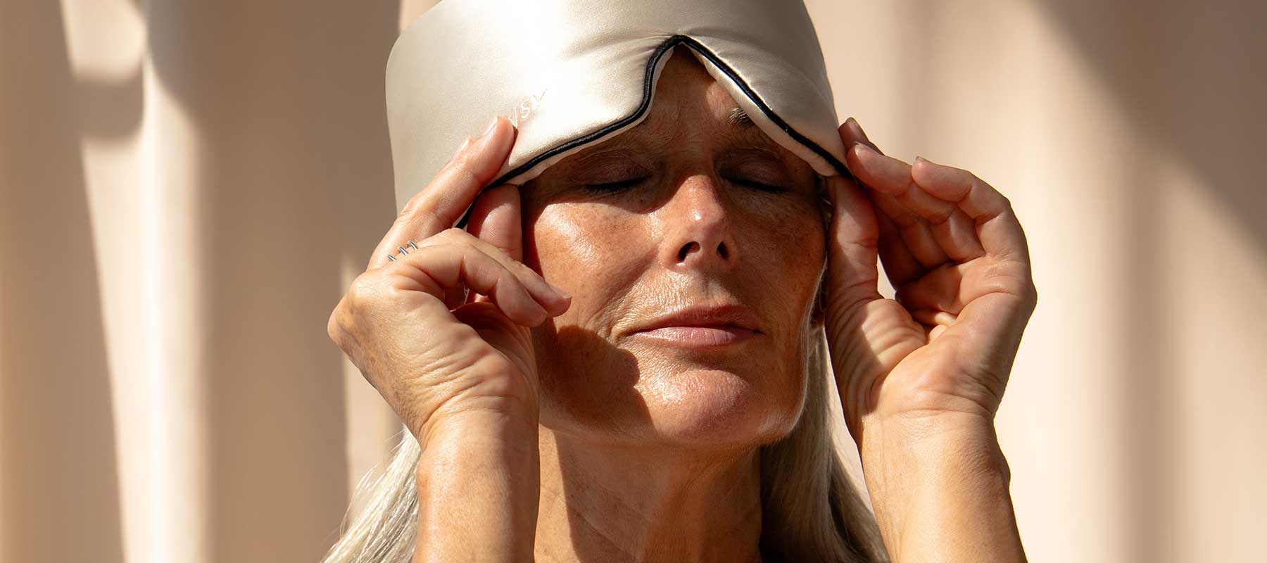 An older woman with long gray hair adjusts a silky sleep mask on her forehead, eyes closed, in soft natural light.