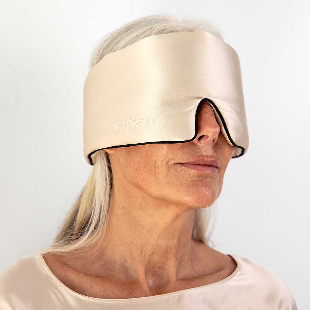 An older woman with long gray hair wears a wide, beige silk sleep mask labeled Drowsy that covers her eyes and ears. She is dressed in a matching satin top and stands against a plain light background.