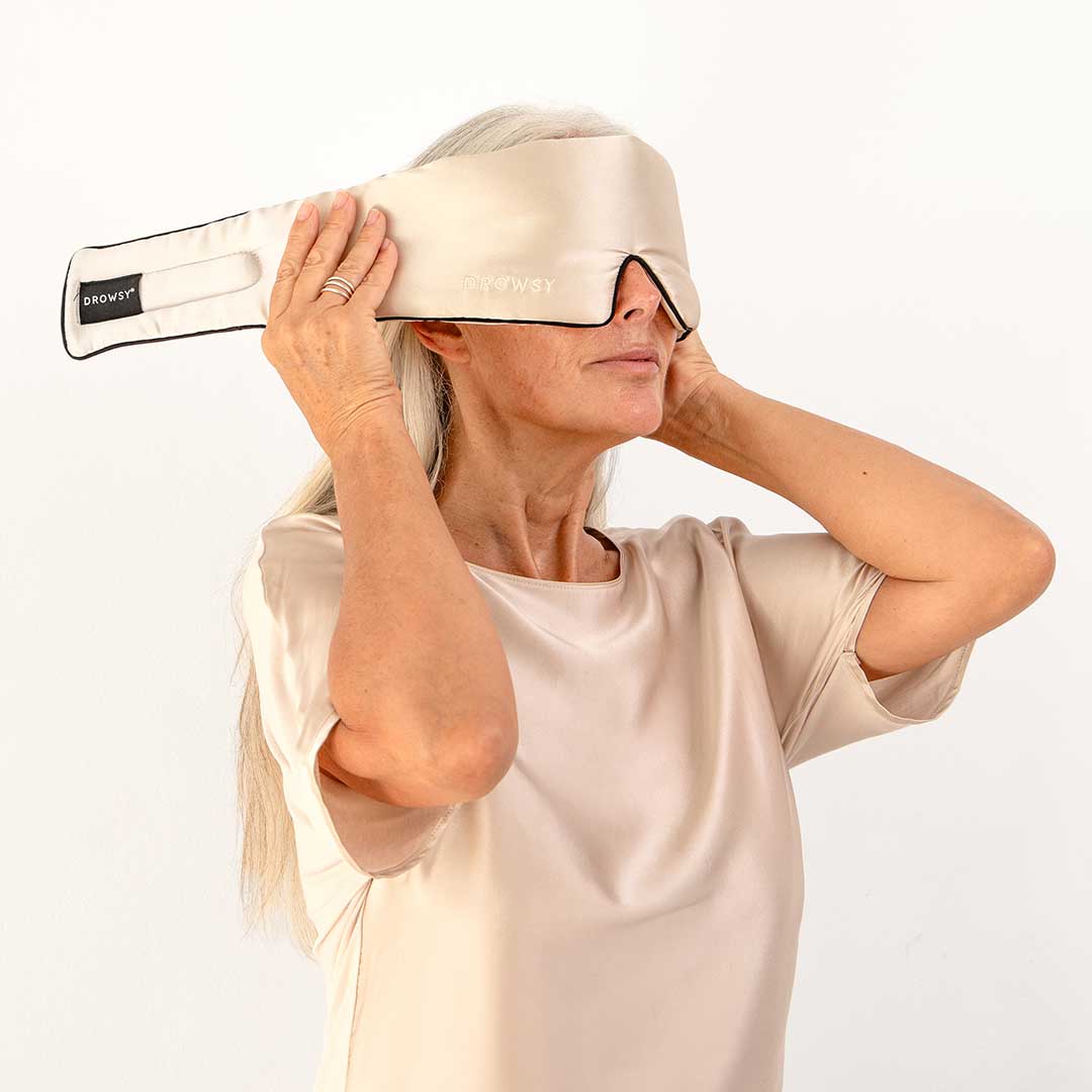 An older woman with long gray hair puts on a beige silk sleep mask while wearing a matching satin short-sleeve top, standing against a plain white background.