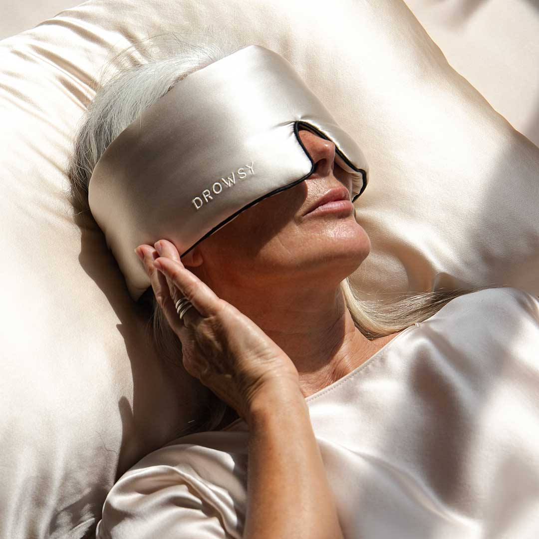 A person with long gray hair lies on a silk pillow, wearing a shiny silk sleep mask labeled DROWSY and matching silk sleepwear, gently touching the edge of the mask.