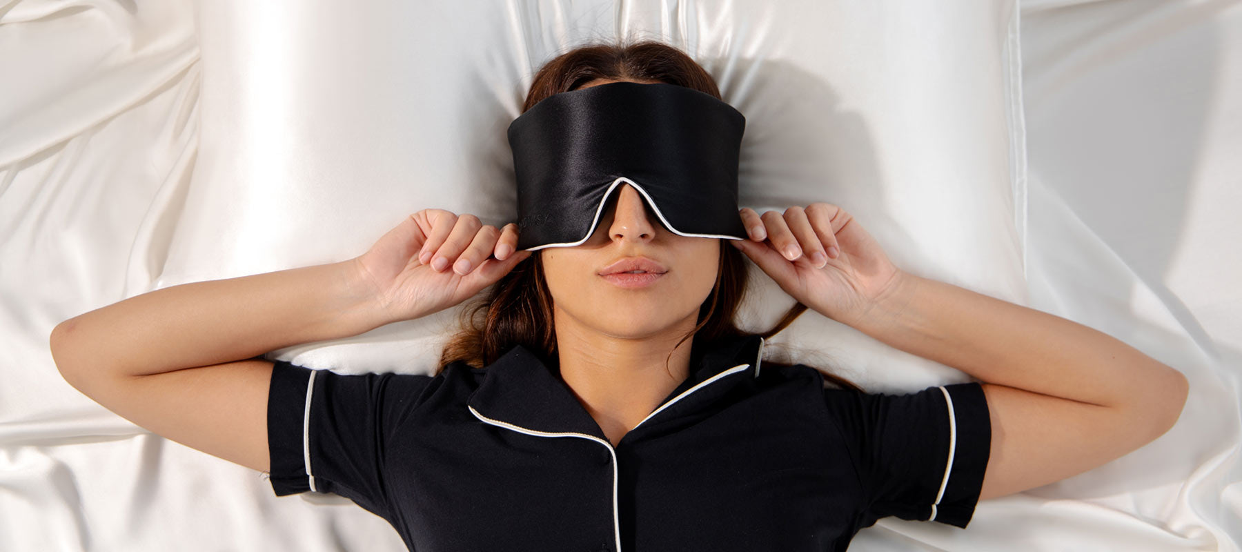A woman lying on a white bed wears a black sleep mask and black pajamas with white trim, holding the mask over her eyes with both hands.