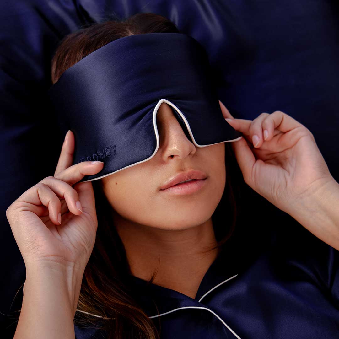A person lying down wears a navy blue sleep mask and navy blue pajamas with white piping, gently adjusting the mask with both hands. The background is also navy blue.
