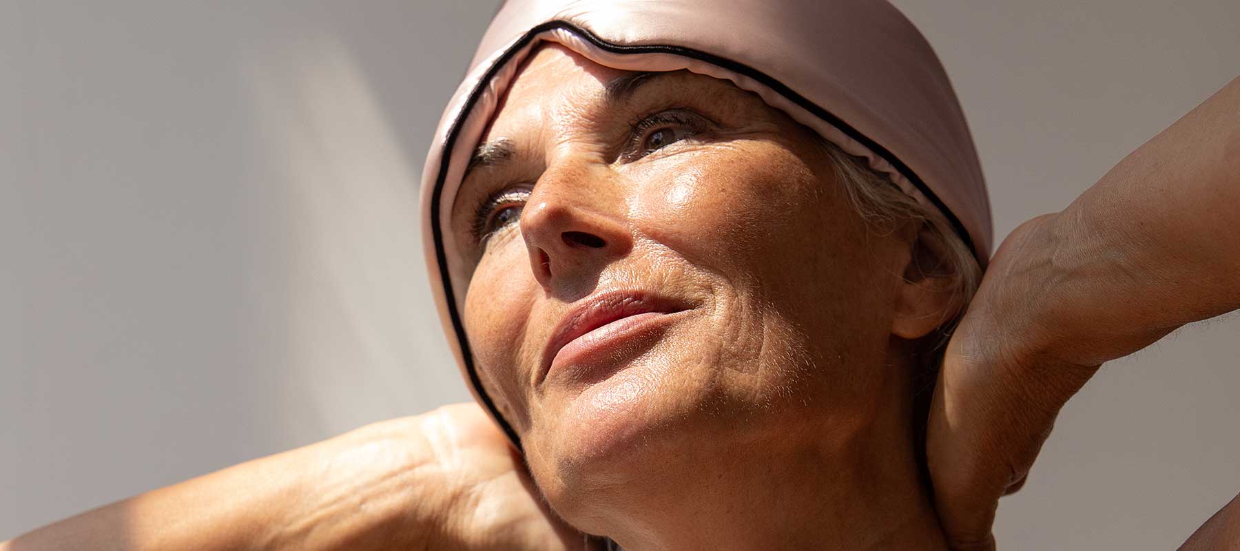 Close-up of an older woman wearing a pink sleep mask pushed up on her forehead, looking content with her hands behind her neck, illuminated by soft natural light.