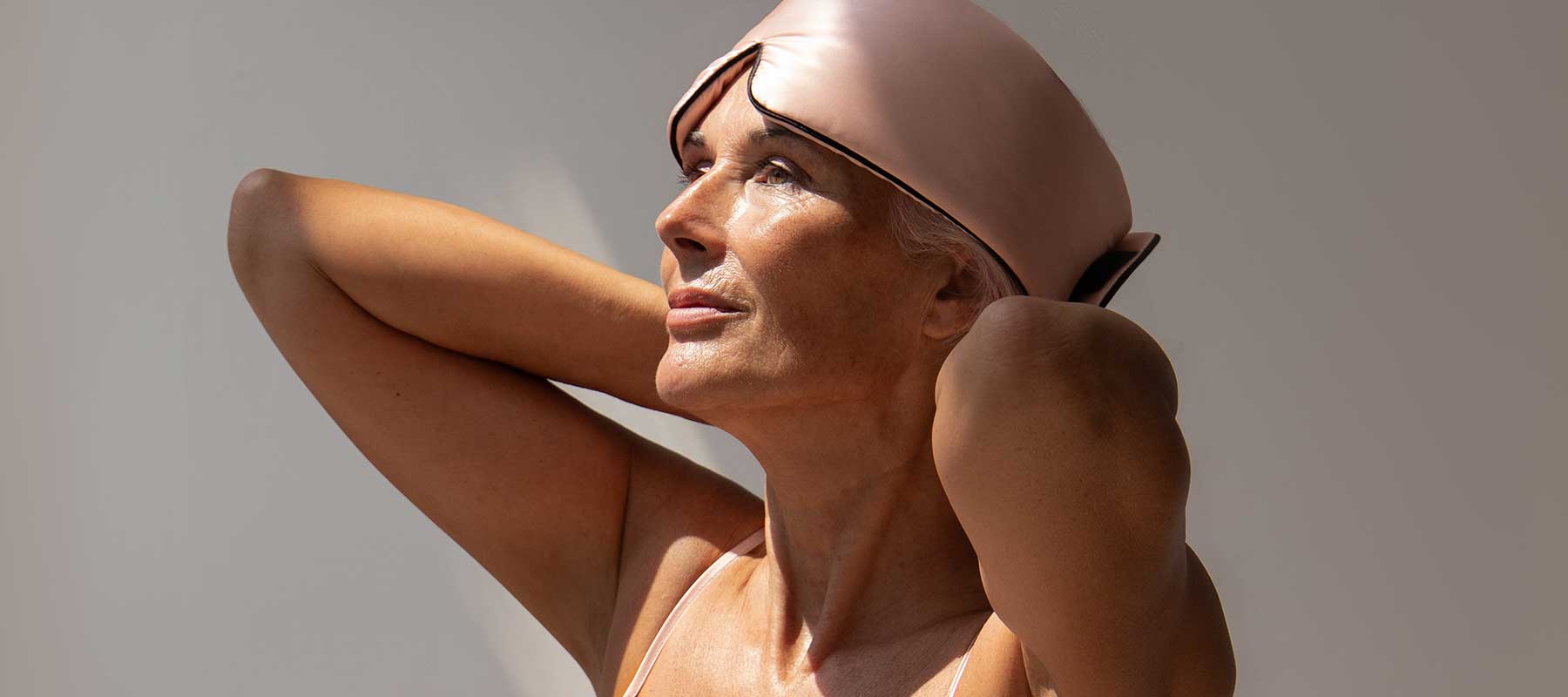A woman with gray hair and glowing skin looks up thoughtfully, wearing a light pink head wrap and a sleeveless top, with her hands behind her head against a neutral background.