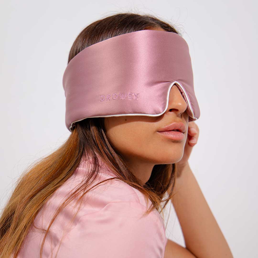 A woman wearing a light pink satin sleep mask with the word DROWSY embroidered on it, sitting with her head tilted slightly to the side and dressed in a matching pink shirt.