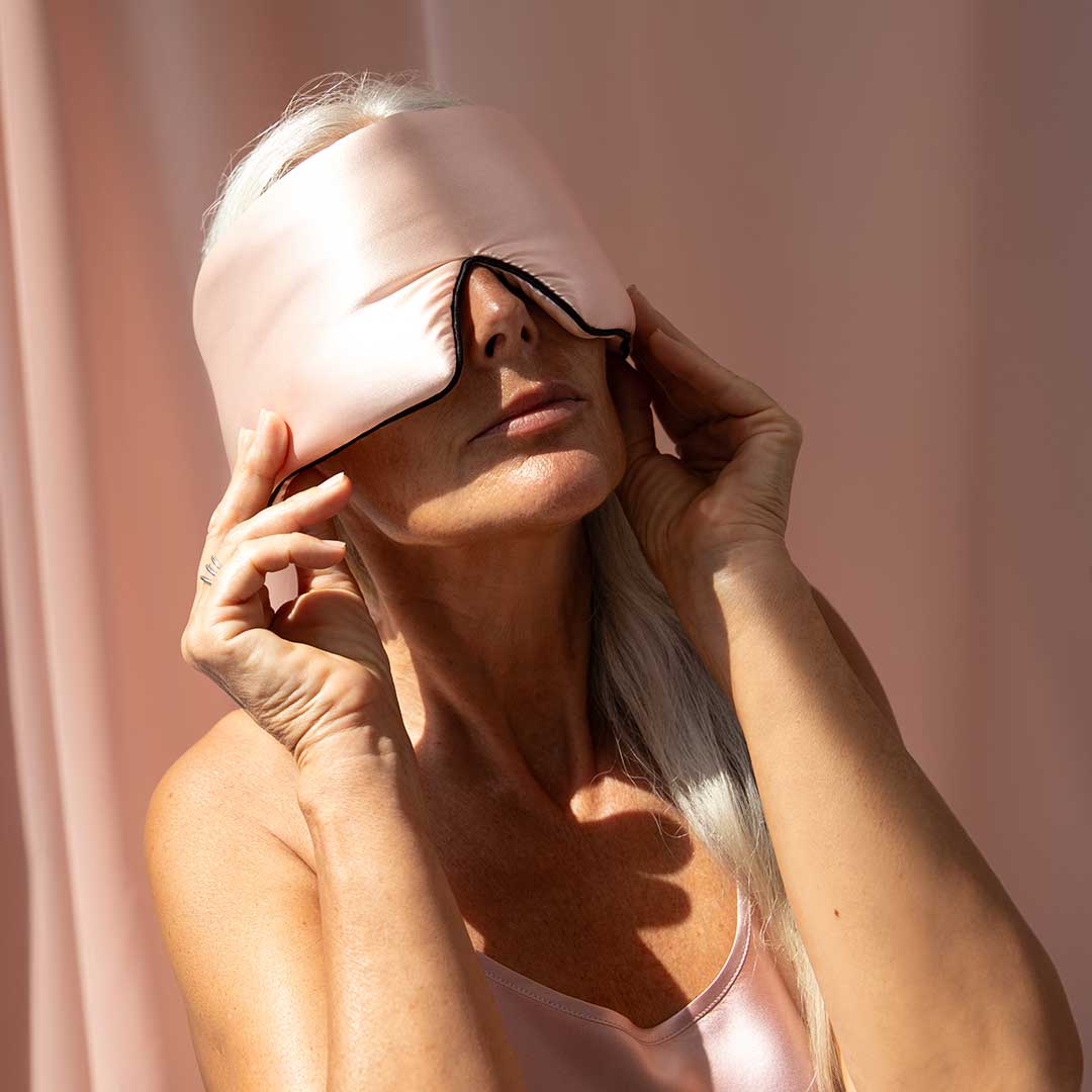A woman with long, silver hair adjusts a large, pink silk sleep mask over her eyes. She is wearing a matching pink satin top, standing against a soft pink background in bright light.