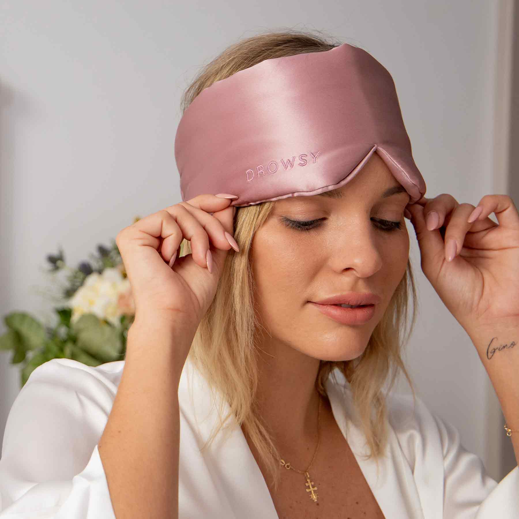 A woman with blonde hair and light makeup adjusts the Damask Rose silk eye mask on her face. She wears a white top, while soft, blurred flowers in the background create a dreamy beauty sleep vibe.