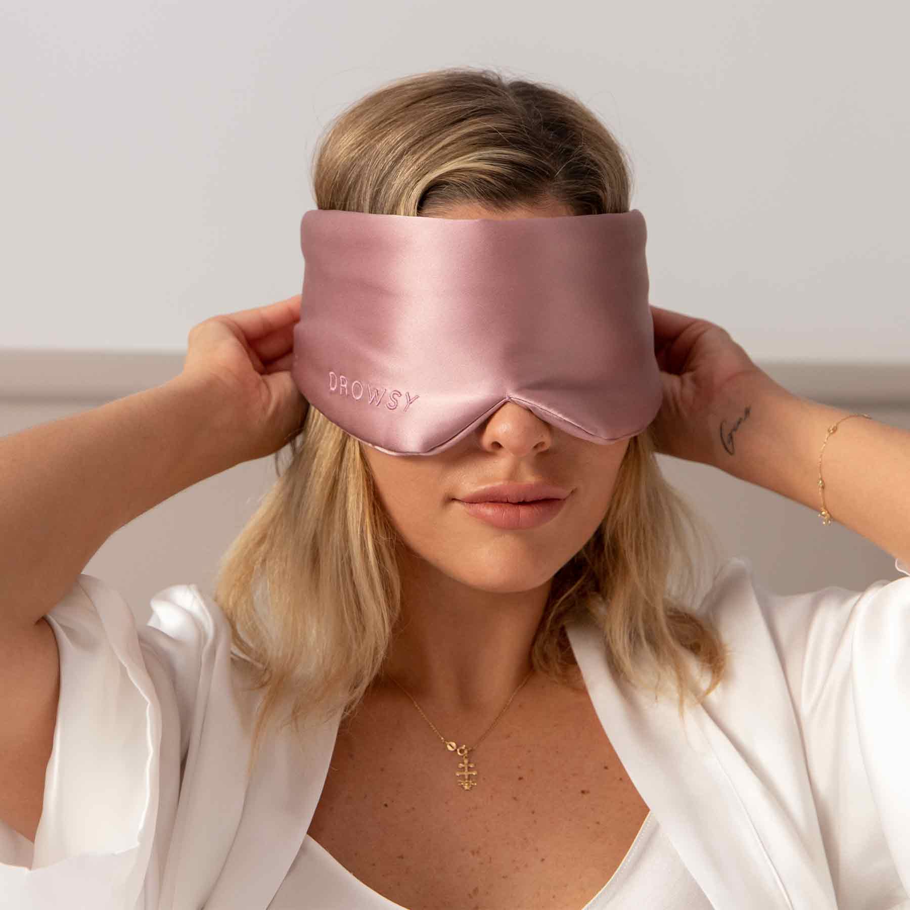 A blonde woman in a white robe wears the large, silky pink Damask Rose eye mask, labeled Drowsy. She holds it in place with both hands against a softly lit, neutral background, creating a soothing beauty sleep vibe.