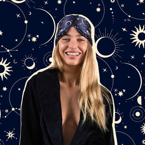 A smiling woman with long blonde hair wears a dark headscarf and robe, embodying beauty sleep with Cosmic Energy against a navy blue background adorned with illustrated stars, moons, and celestial patterns.