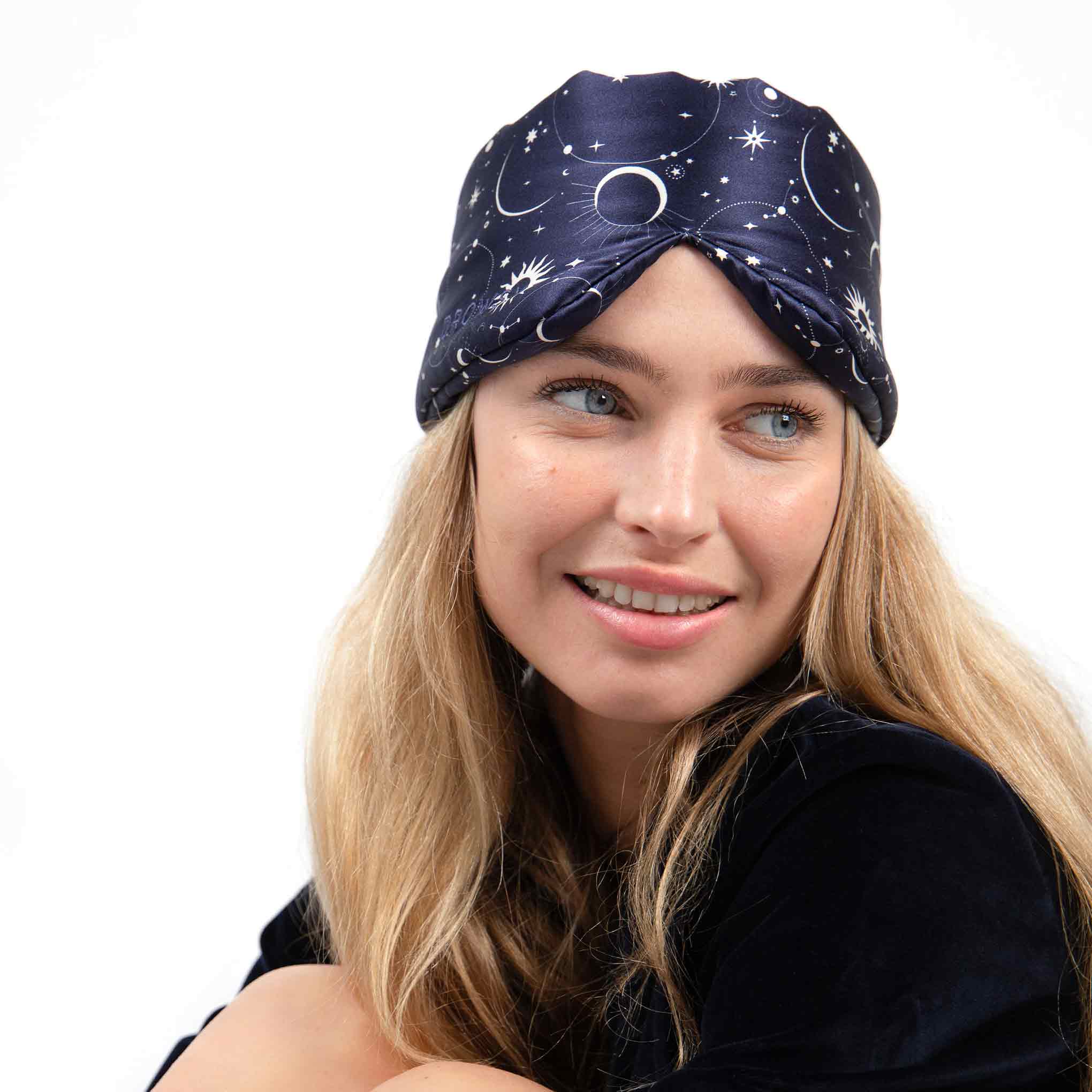 A woman with long blonde hair wears the Cosmic Energy dark turban-style headband featuring a white moon and stars pattern, smiling and looking to the side against a plain white background, radiating beauty sleep vibes.