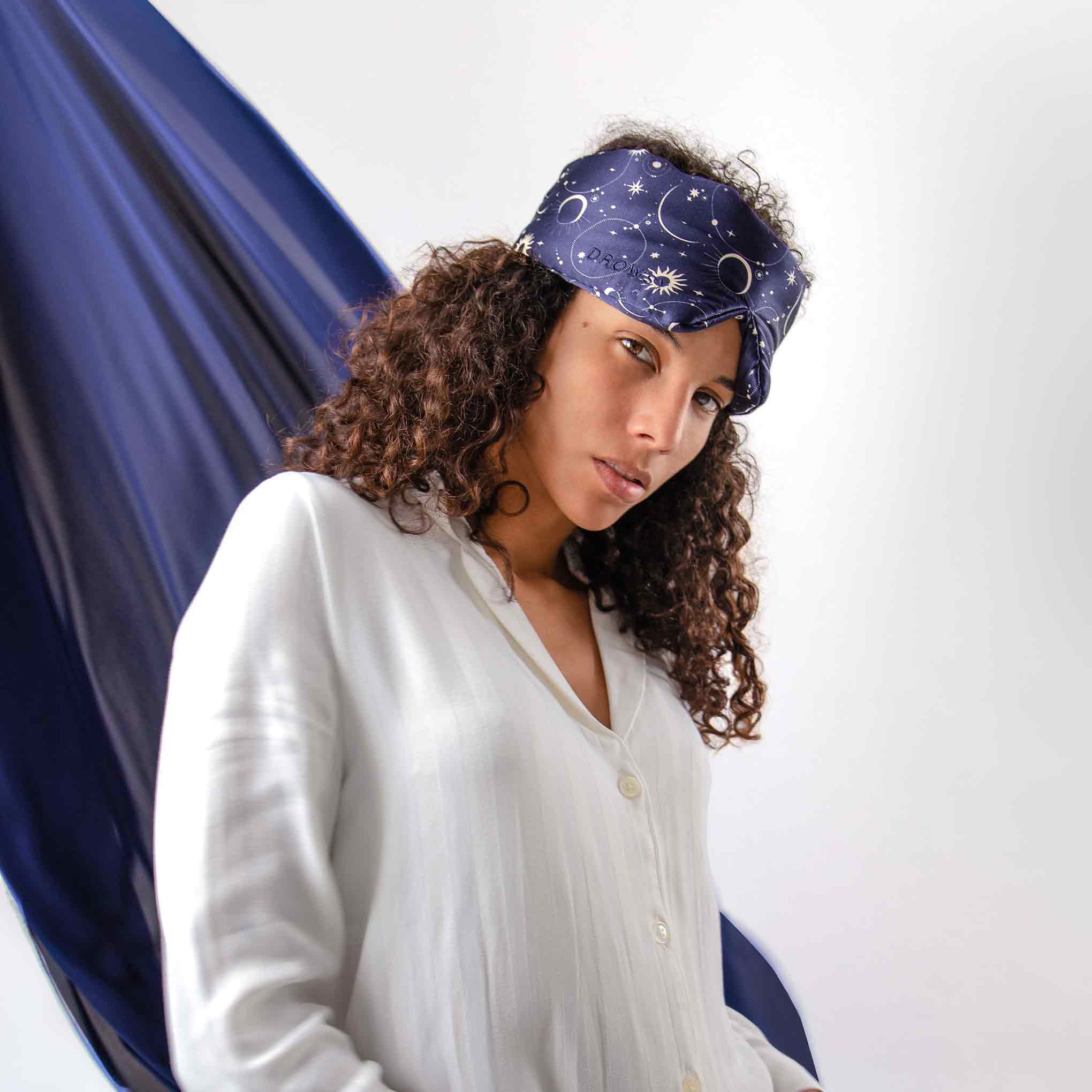 A person with curly hair wears a white button-up and the Cosmic Energy navy blue silk eye mask featuring stars and moons, standing before a white backdrop with flowing dark blue fabric.