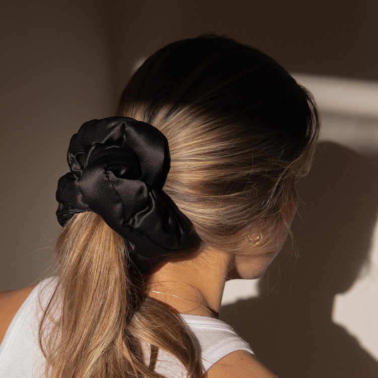 A woman with light brown hair in a low ponytail, secured with drowsysleepco’s Sensual Sleep Bundle - Black Jade scrunchie, wears a white sleeveless top and is shown from behind in soft natural light.