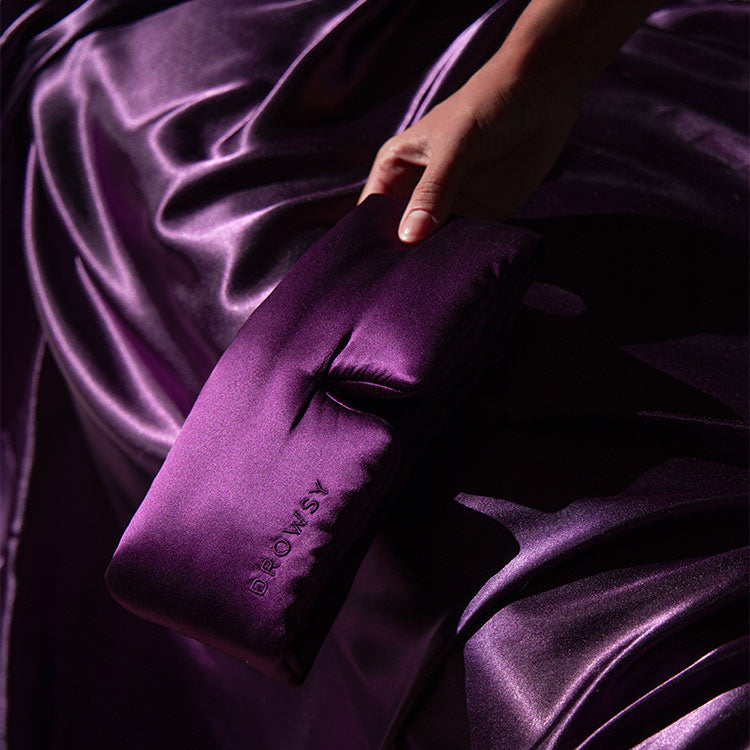 Hand reaching for Drowsy silk sleep mask on purple coloured silk