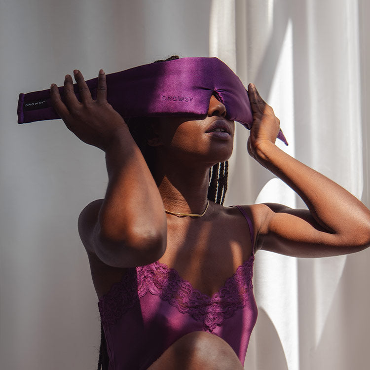 Model putting on Drowsy Purple Martini Silk Sleep Mask with sun shining on her