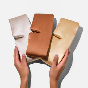 Two hands hold the Besties Pack—three silky sleep masks in ivory, copper, and gold—against a white background. Each mask features “DROWSY” embroidered on it.