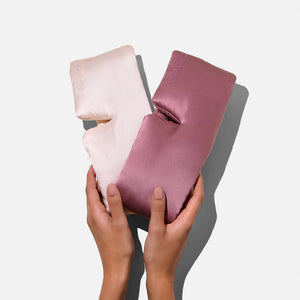 Two hands hold two silky Lovers Pack eye masks—one light pink, one mauve—against a plain light gray background.