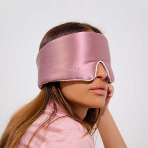 A woman wears the Damask Rose - Pipe Dreams Edition, a luxurious wide pink mulberry silk eye mask embroidered with DROWSY, partially covering her face and standing out against a plain light background.