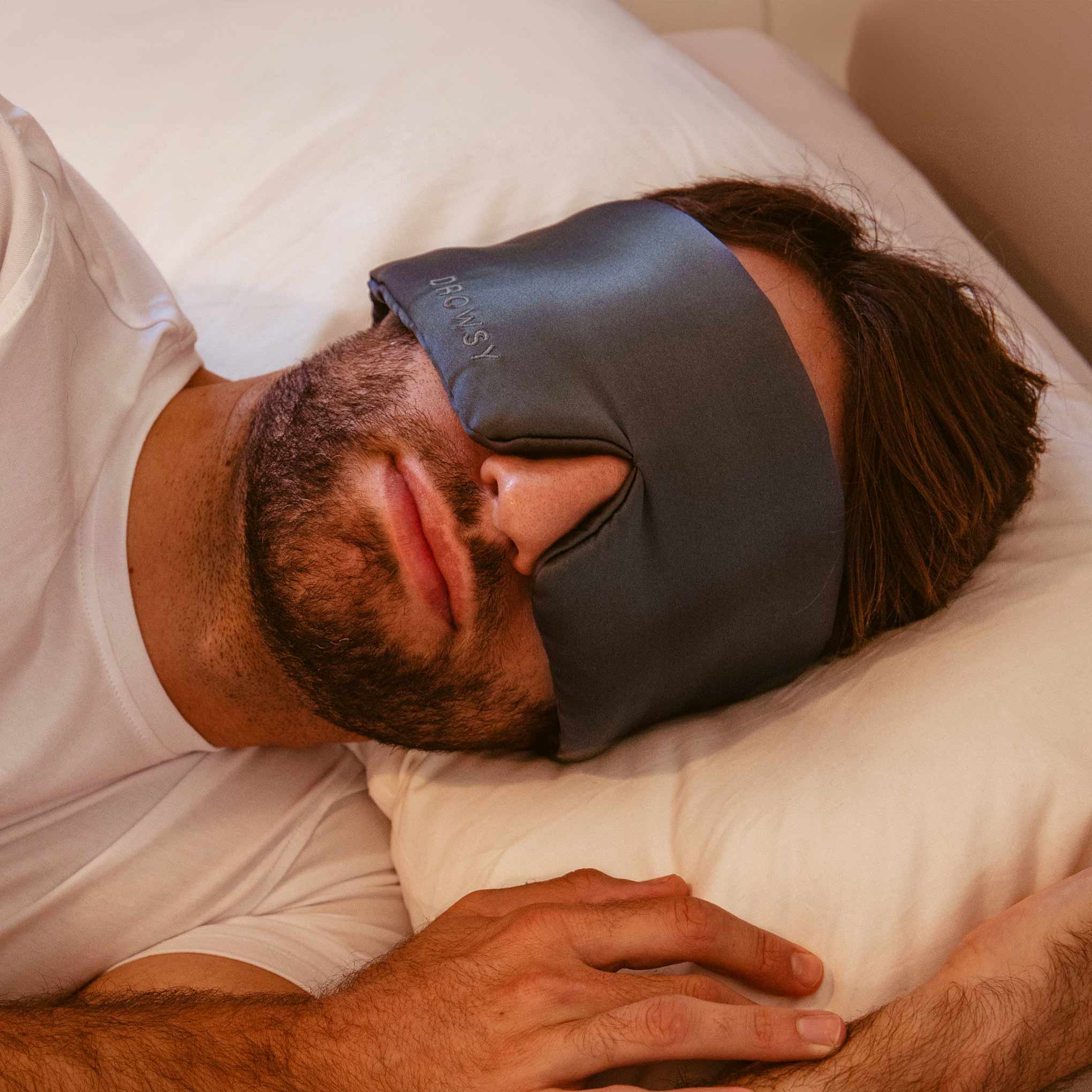 A bearded man in a white t-shirt lies on a bed, resting on a white pillow while wearing the drowsysleepco Moonlight Shadow blackout eye mask with nose gap for perfect beauty sleep.
