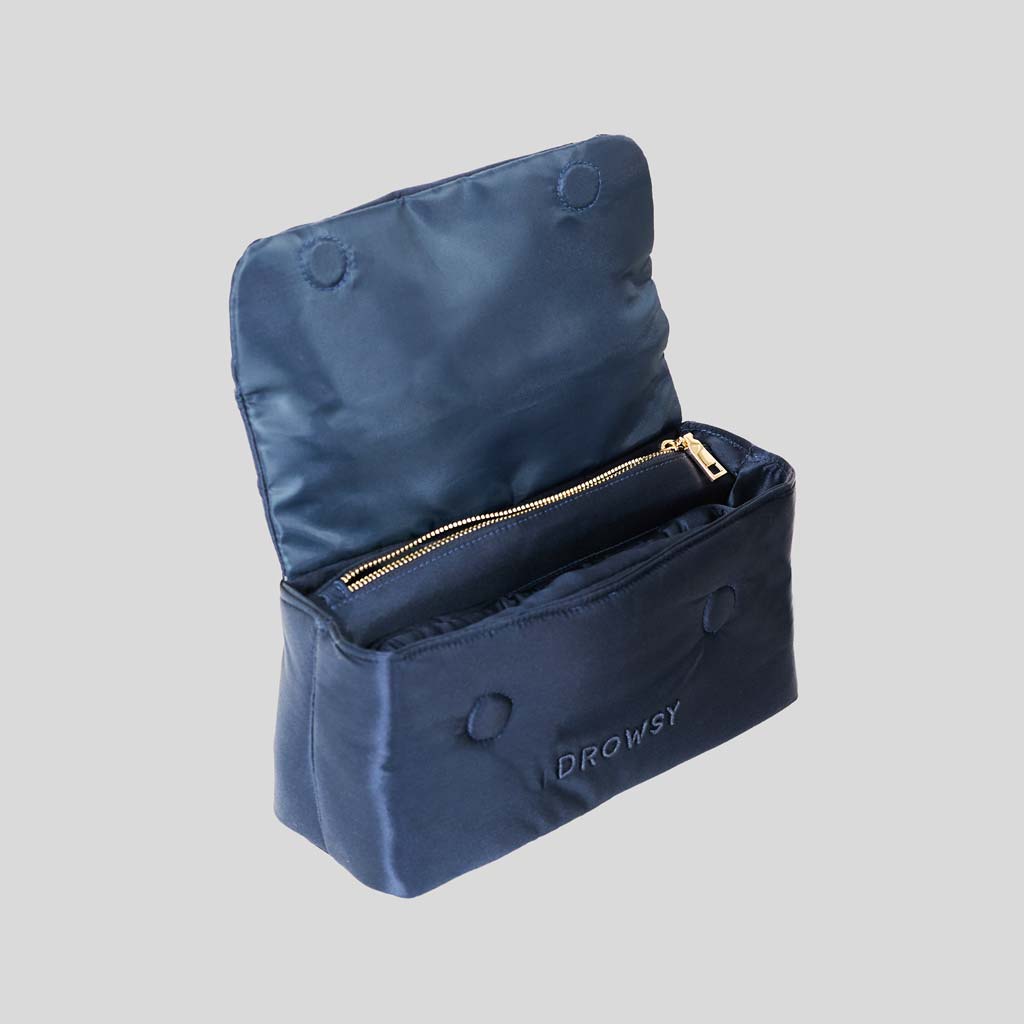 The Drowsy Silk Necessaire in Midnight Blue is shown open, revealing a zippered compartment and lined interior with buttons—perfect for elevating your beauty routine. DROWSY is embroidered on the front against a plain gray background.