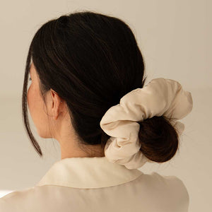 A woman with dark hair in a low bun, secured with the Dusty Gold - Padded Scrunchie. She wears a cream collared shirt and is photographed from behind against a neutral background.