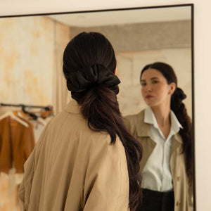 A woman with long dark hair tied in a Black Jade - Padded Scrunchie gazes at her reflection in a mirror. She wears a beige coat over a white blouse, with clothes hanging in the background.