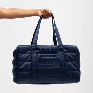 A manicured hand holds the Drowsy Silk Weekender - Midnight Blue, a spacious quilted duffel bag with DROWSY embroidered on the front, displayed against a simple light background.