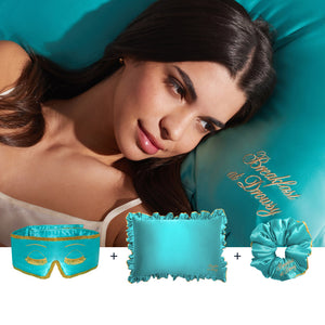 A woman with long dark hair lies on a turquoise satin pillow embroidered with "Breakfast at Draper," alongside a matching sleep mask, ruffled pillowcase, and scrunchie—all part of the Hollywood Experience Bundle.